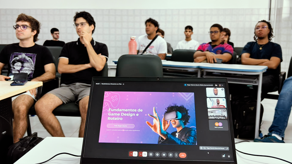 Foto tirada por trás de uma tela de notebook mostrando uma videoconferência com o slide "Fundamentos de Game Design e Roteiro". Ao fundo, vários jovens estudantes estão sentados em carteiras de uma sala de aula prestando atenção à aula.